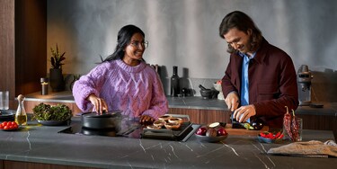 Woman in pink sweater cooking on hob next to man in red jacket cutting vegetables